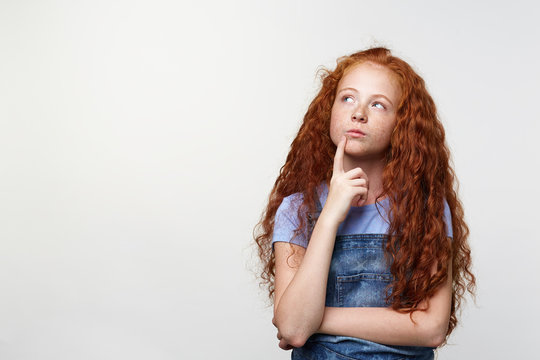 Photo of thinking cute freckles little girl with ginger hair, looks serious and doubt, touches chin, looks away over white background with copy space on the left side.