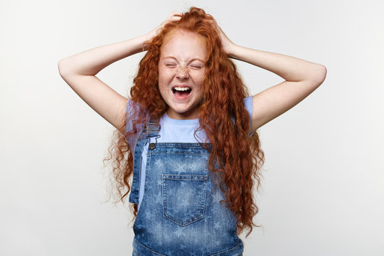 Photo of sceaming freckles little girl with ginger hair, with raised hands, holding head, stands over white wall with closed eyes and wide open mouth, looks unhappy.
