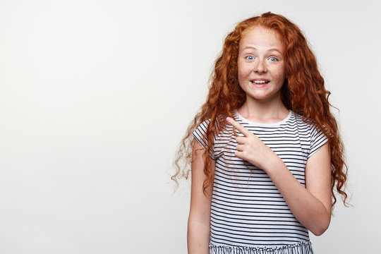 Portrait of happy amazed little girl with ginger hair and freckles, wants to draw you attention to the copy space on the left side and points with fingers, stands over white wall and broadly smiling.