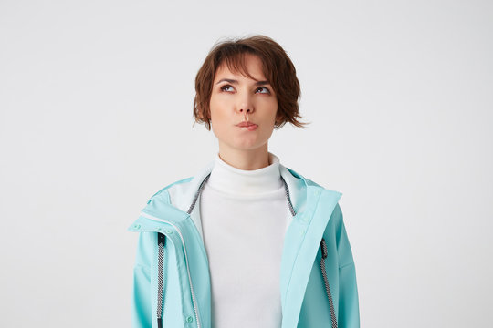 Photo of young wishful short-haired lady in white golf and light blue rain coat, bites lips and looks up, stands over white background.