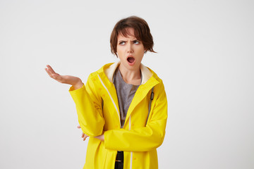 Shocked short-haired young woman in yellow rain coat, with wide open mouth and outraged expression,...