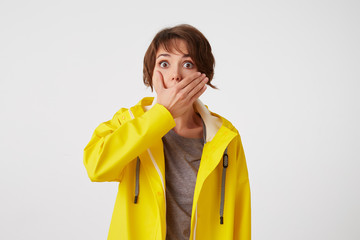 Portrait of short-haired curly woman in yellow rain coat, hears unbelievable news , covered mouth with hand, stands over white wall with wide open eyes with surprised expression.