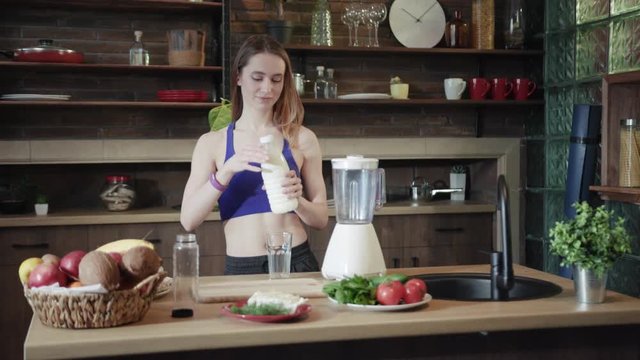 Beautiful fit young Caucasian woman in sportswear pours out milk in glass, drinks it with pleasure and smiles happily on modern kitchen background. Healthy nutrition, body caring, active lifestyle