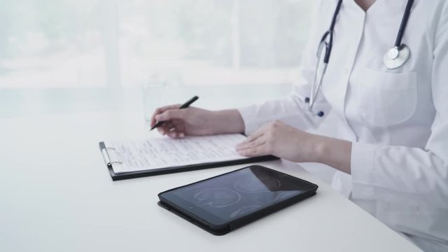 Female caucasian doctor using tablet and writing diagnosis in medical form and documents. Close up 4K