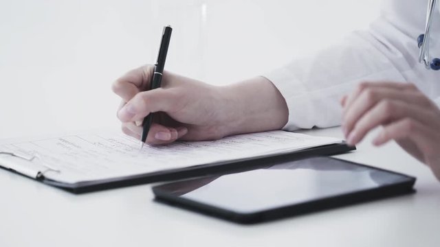 Female caucasian doctor using tablet and writing diagnosis in medical form and documents. Close up 4K