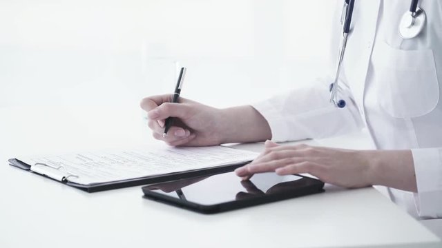 Female caucasian doctor using tablet and writing diagnosis in medical form and documents. Close up 4K