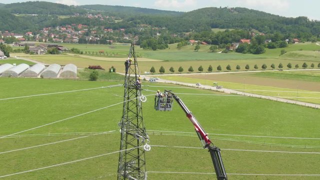 AERIAL Workers standing inside telescopic boom lift and attaching high voltage wires to a power tower in the scenic countryside. Maintenance crew working on electricity pylon next to a regional road.