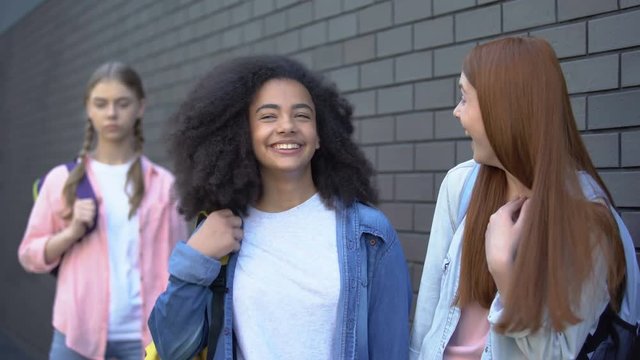 Two schoolgirls making fun of female student standing aside, spreading rumors