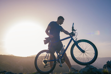 Naklejka na ściany i meble Mountain biker holding his bike on a rough cliff terrain on a sunset.