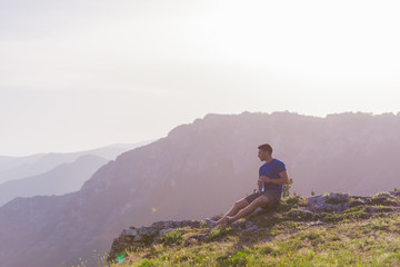 Handsome athlete sitting at a rocky peak while looking at the breathtaking mountain line and a beautiful lake while wearing a blue shirt and grey shorts.