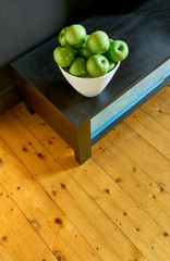 A group of green Granny Smith cooking apples in a white bowl on a brown wooden bench  and a vibrant wooden floor from above.