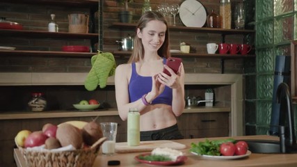 Cheerful attractive young sportswoman using phone over cozy kitchen background. Browsing social networks, surfing the internet, fitness app. Technology and healthy lifestyle concept