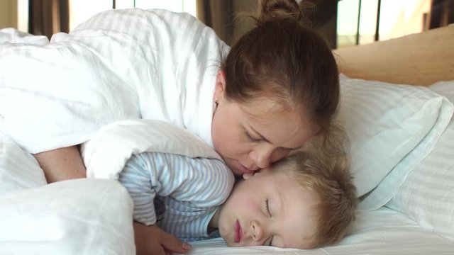 A caring mother kisses her sleeping son in the morning in bed, she wakes him up. A high resolution. Mom and son are sleeping in a white bed in the morning, they woke up.