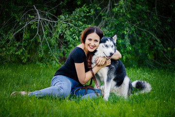 Naklejka na ściany i meble A beautiful girl is sitting on the lawn with her dog. Siberian husky dog with blue eyes. Bright green trees and grass are in the background. Friendship between man and animals.