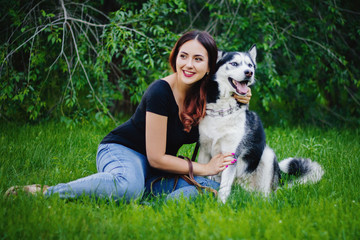 Naklejka na ściany i meble A beautiful girl is sitting on the lawn with her dog. Siberian husky dog with blue eyes. Bright green trees and grass are in the background. Friendship between man and animals.