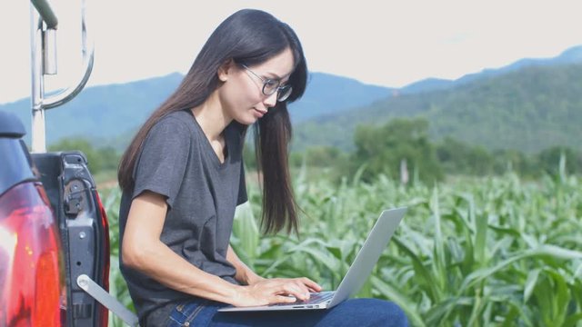 Young happy girl sitting and working on computer. Asian woman near car in meadow holding laptop. Person working outdoor. 