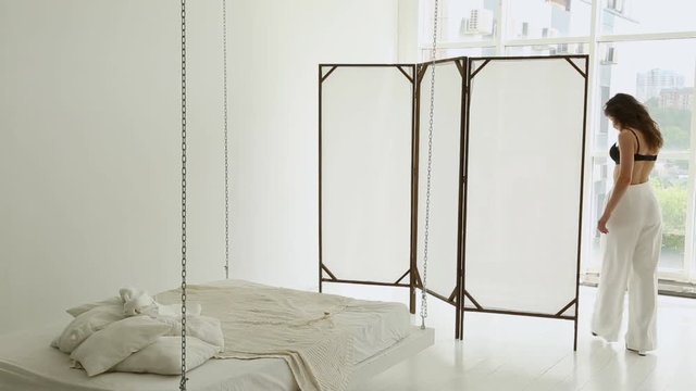a brunette girl in white trousers and black lingerie and shoes walks over the white room, next to a hanging bed, large windows to the floor, removes the bra behind the screen and hangs it.