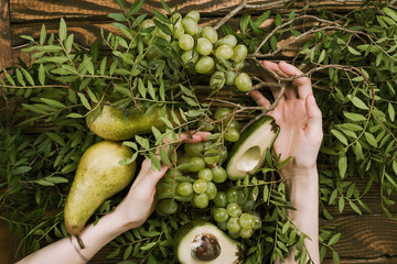 Flat lay of woman's hands holding grapes, pears and avocados with pistachio branches on wooden table