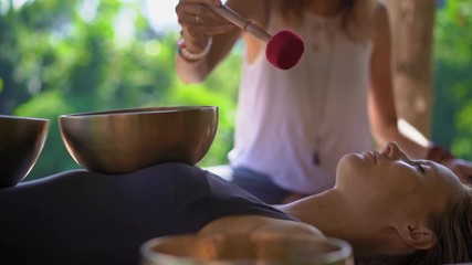 Superslowmotion shot of a woman master of Asian sacred medicine performs Tibetan bowls healing ritual for a client young woman. Meditation with Tibetan singing bowls. They are in a gazebo for