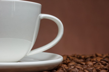 White cup of coffee close-up on coffee beans side view on brown background.