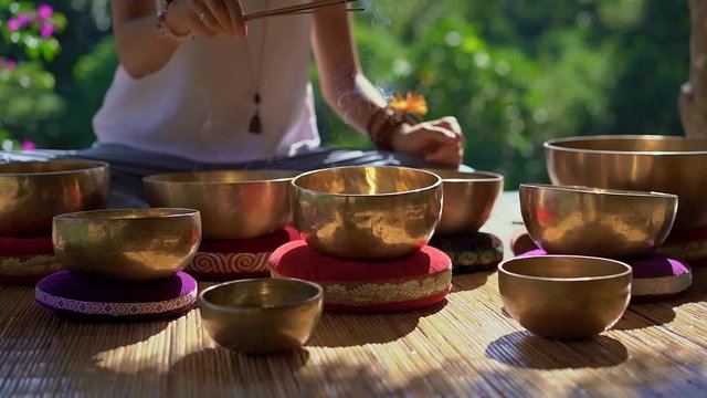 Superslowmotion shot of a woman master of Asian sacred medicine performs Tibetan bowls healing ritual. Meditation with Tibetan singing bowls. She sits in a gazebo for meditation with a beautiful
