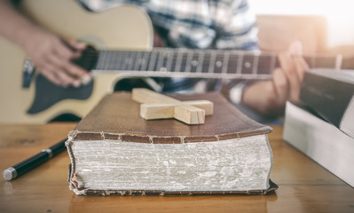 Close up the old Bible and wooden cross with young male playing guitar acoustic at background. Christian worship God concept.