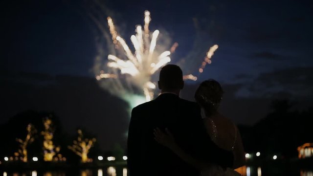 Happy hugging bride and groom watching beautiful colorful fireworks night sky