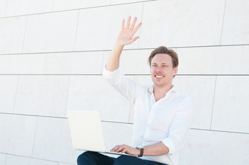 Happy young man waving to his friend outdoors. Positive digital nomad with laptop bringing...