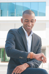 Portrait of smiling young businessman looking at watch. African American manager sitting at office building checking time. Punctuality concept