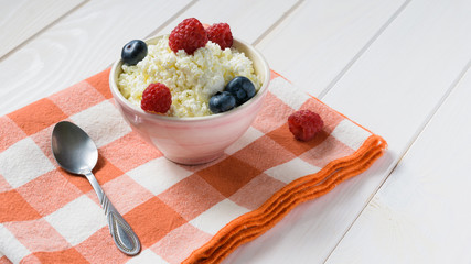 bowl of cottage cheese with raspberries and blueberriesand a spoon on the red plaid litter on a white wooden table