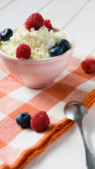bowl of cottage cheese with raspberries and blueberriesand a spoon on the red plaid litter on a white wooden table