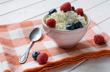 bowl of cottage cheese with raspberries and blueberriesand a spoon on the red plaid litter on a white wooden table