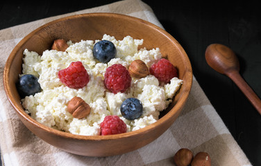 wooden bowl with cottage cheese, blueberries, raspberries and hazelnuts on a black wooden table