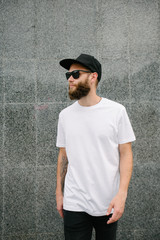 Hipster handsome male model with beard wearing white blank t-shirt and a baseball cap with space for your logo or design in casual urban style