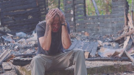 Portrait of a sad man on the background of a burned house, after fire or artillery attack. Consequences of fire disaster accident. Ruins after fire disaster, loss and despair concept.