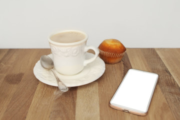Mock up image of white mobile phone with empty white screen, cup of coffee and cupcake on wooden table
