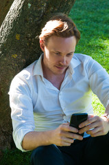 Young businessman using mobile internet while resting in park. Introspective man in white shirt reading message on phone. Lifestyle concept