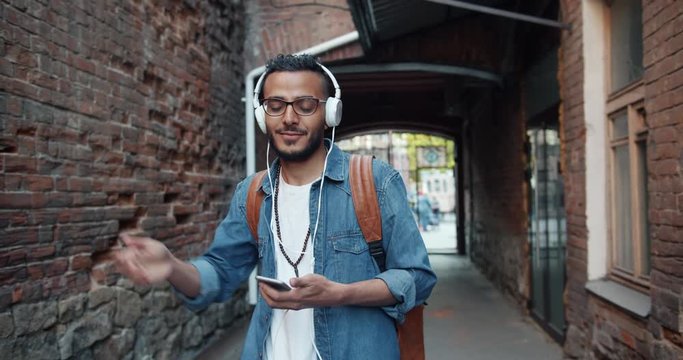 Slow motion of young Arabian hipster enjoying music in headphones dancing outdoors having fun holding smartphone. Youth and modern gadgets concept.