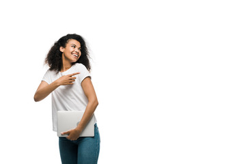 happy young african american woman pointing with finger while holding laptop isolated on white