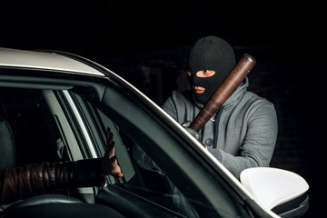 robber in a Balaclava tries to break the car glass with a baseball bat.