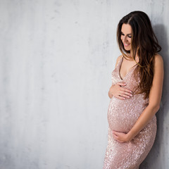 portrait of happy pregnant woman in shiny evening dress looking at her tummy over gray wall...