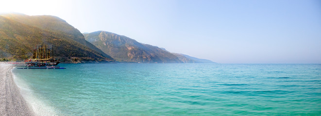 Naklejka premium Secluded beach surrounded by valley cliffs in a tranquil bay with turquoise water and sailing boats at sunrise, Oludeniz, Turkey panoramic
