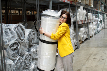 Girl with glasses takes a big and heavy mattress in the store. Wholesale and purchase. Self-service warehouse. Selection of products