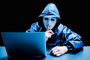 Anonymous computer hacker in white mask and hoodie. Obscured dark face making silence gesture try...