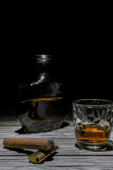 A photo of a glass of whiskey, a cigar with a lighter and a bottle of alcohol are on the old textured wooden table. A very deep dark background behind.