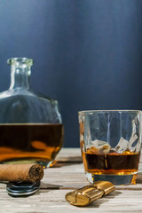 Vertical photo of a glass of whiskey, a cigar with a lighter, guillotine and a bottle of alcohol are on the old textured wooden table.