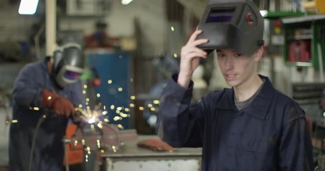 4K Young welder removing visor & smiling at camera with coworker in background. Slow motion