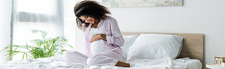 panoramic shot of attractive pregnant african american woman looking at belly and sitting on bed © LIGHTFIELD STUDIOS