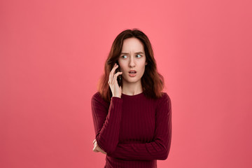 Portrait of a beautiful brunette girl standing on a dark pink background and looking worried while speaking on the phone.