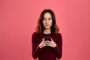 Portrait of a pretty brunette girl standing on a dark pink background and texting on the phone.